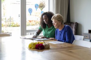 A compassionate caregiver in a green uniform assisting an elderly woman in a blue sweater with reading or paperwork at a wooden table. The setting is a bright and cozy home environment with knitting supplies on the table, suggesting a warm and engaging atmosphere.