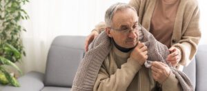 A caring woman placing a warm knitted blanket over an elderly man with glasses, sitting on a couch in a cozy home environment. The senior man wears a beige sweater and looks thoughtful, while the woman provides comfort and warmth.