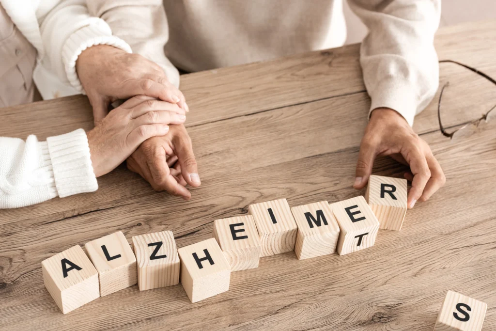 Close-up of elderly couple holding hands with wooden blocks spelling Alzheimer’s on a table.