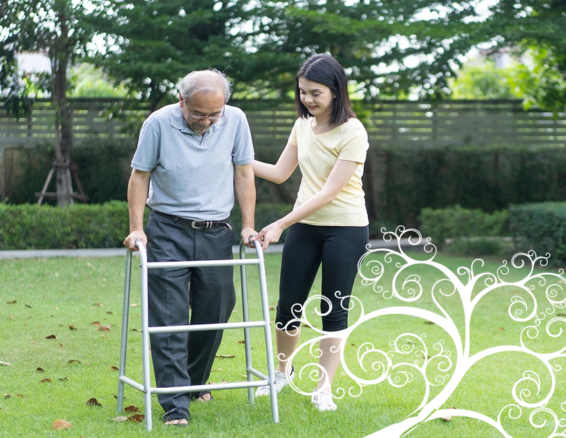 A young woman assists an older man using a walker on a grassy lawn. Both appear focused and supportive, surrounded by green trees, conveying care.