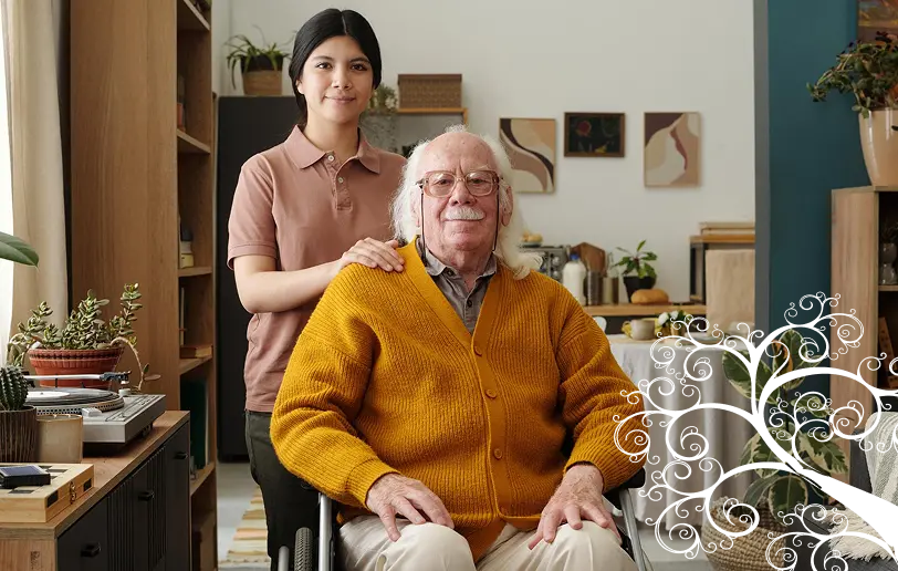 A caregiver standing behind an elderly man in a wheelchair in a warm home interior with a decorative tree illustration
